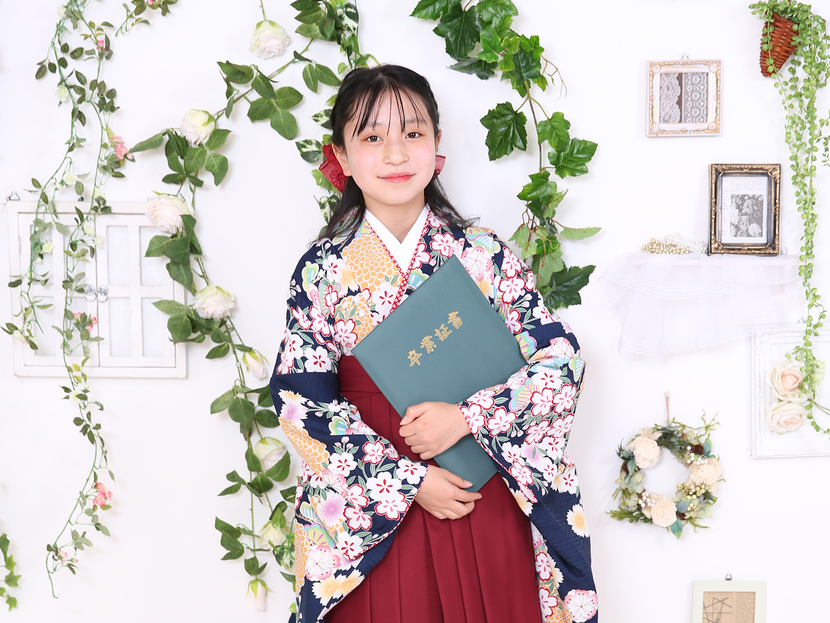 花柄の着物を着た少女が笑顔で卒業証書のような冊子を持っている様子。緑の植物と白い花で飾られた背景の前で、卒業や入学式を記念する和装姿の記念写真。