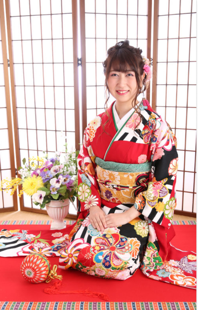 障子の前で座る笑顔の女性。赤い振袖に華やかな模様と青い帯を合わせた姿で、傍らには花のアレンジメントが飾られた和の空間での成人式記念写真。