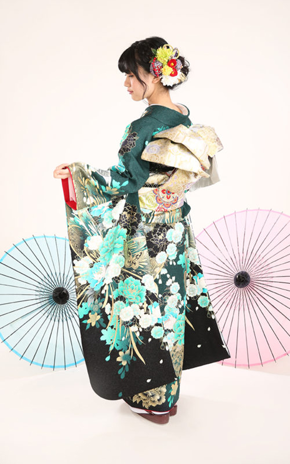 深緑色の地に青や水色の花柄が映える振袖を着た女性の後ろ姿。華やかな髪飾りを付け、背景には和傘が配置された成人式の記念写真。
