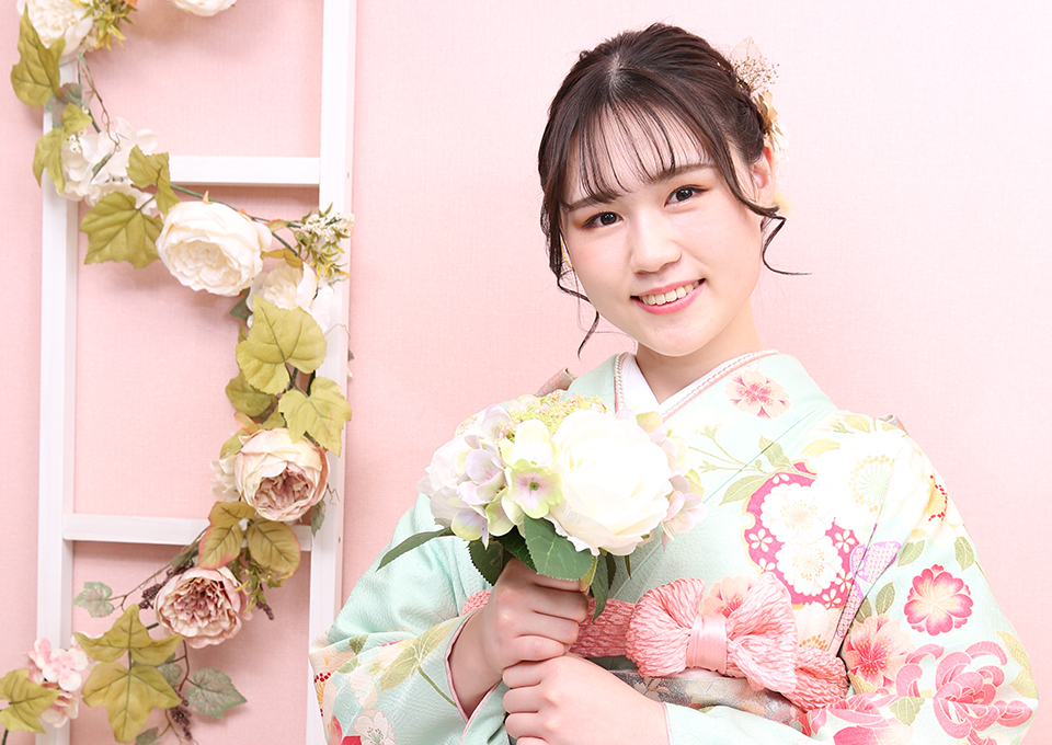 赤いカーテンを背景に、赤い振袖姿の若い女性がカラフルな花束を持って微笑んでいる様子。髪には華やかな花の髪飾りをつけた成人式の記念撮影。