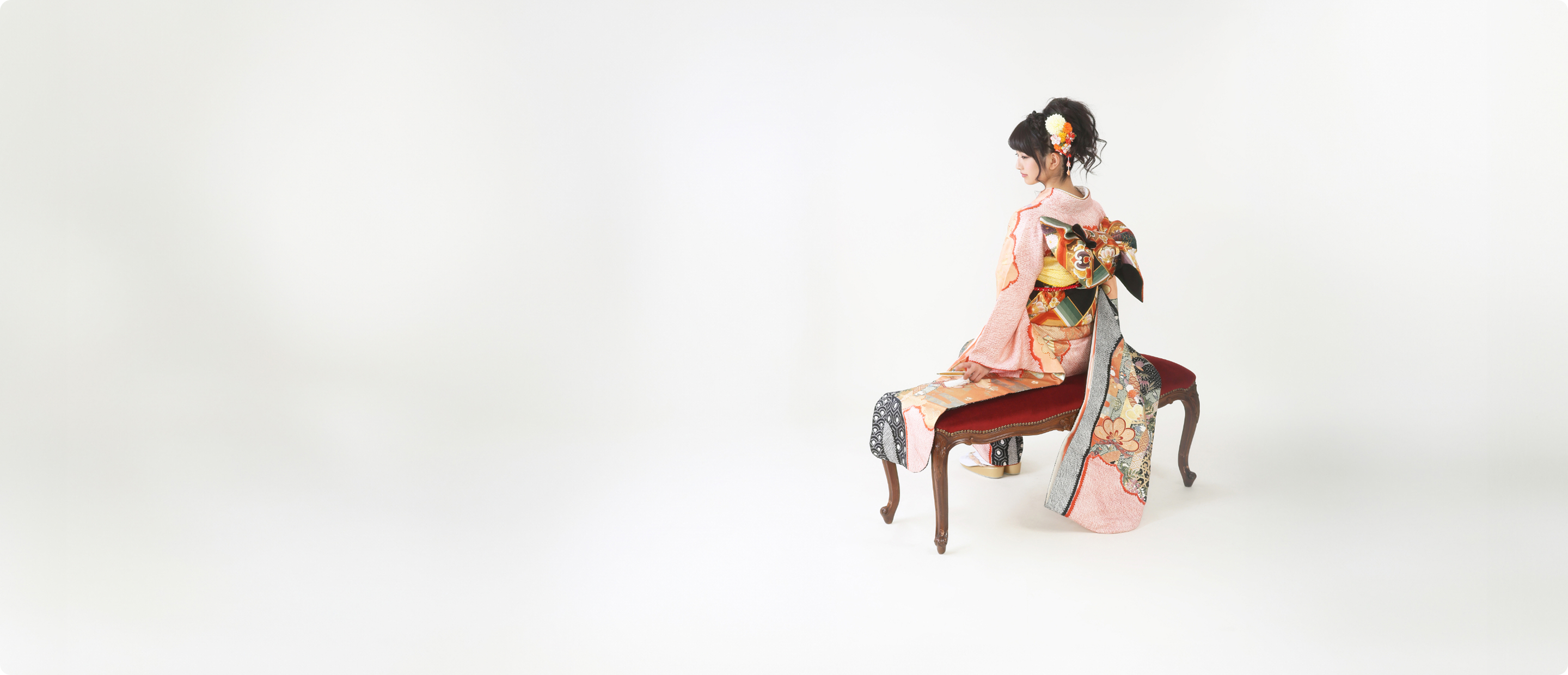 白い背景の前で、ピンク色の着物を着た若い女性が木製の椅子に座っている姿。華やかな帯と優美な着付けが特徴的な成人式や節目の記念写真。